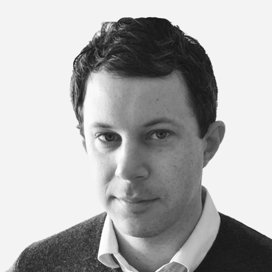 A black and white portrait of a man with short, dark hair. He is wearing a collared shirt and a sweater. His expression is neutral, and he is looking directly at the camera. The background is plain and light-colored.