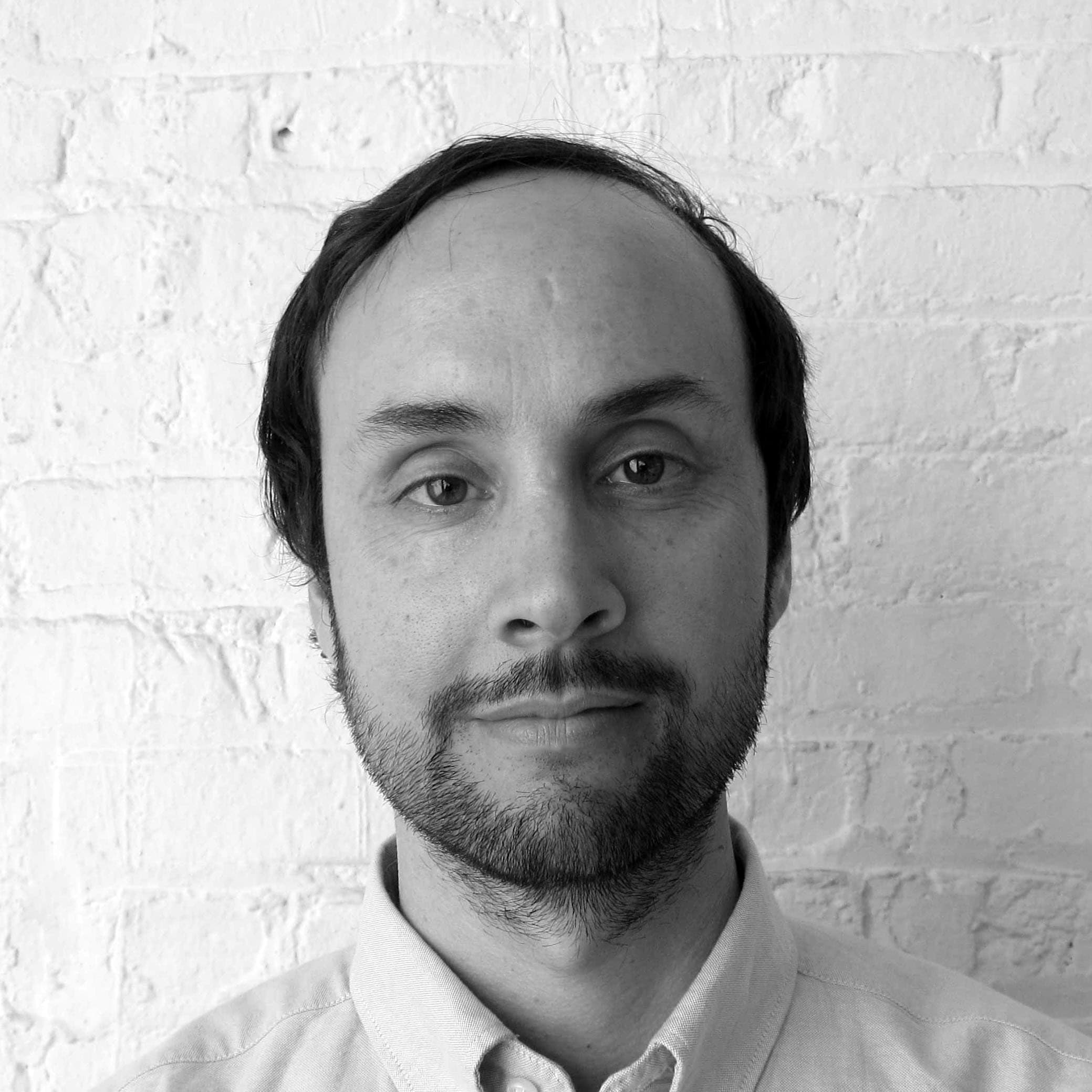 A black and white portrait of a man with short dark hair, a receding hairline, and a beard. He is wearing a light-colored shirt and standing in front of a textured brick wall, looking at the camera.