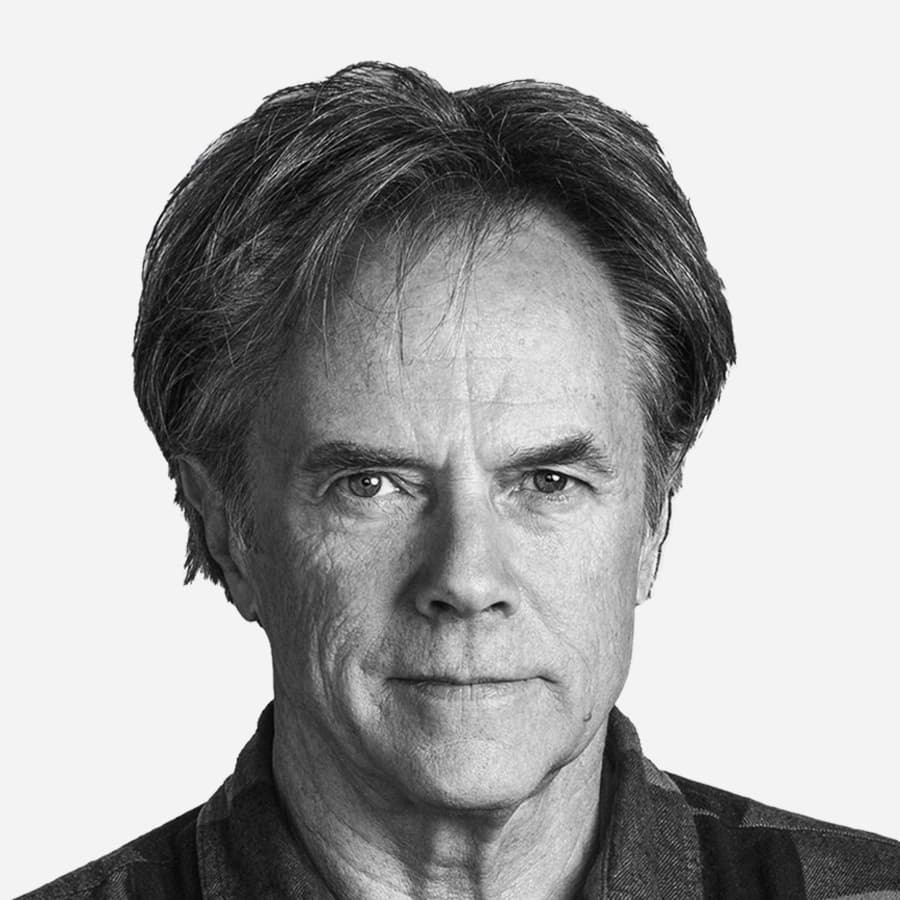 A grayscale photo shows an older man with short, slightly tousled hair, and a serious expression. He is facing forward, and the image captures his face and upper shoulders against a plain background. He is wearing a dark collared shirt.