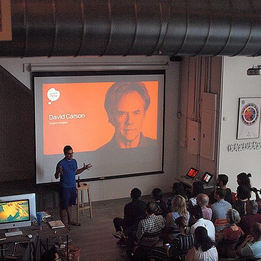 A person stands in front of an audience giving a presentation. The screen behind them displays a large portrait and the name "David Carson" with the title "Graphic Designer" on an orange background. The audience is seated and attentively watching the presentation.