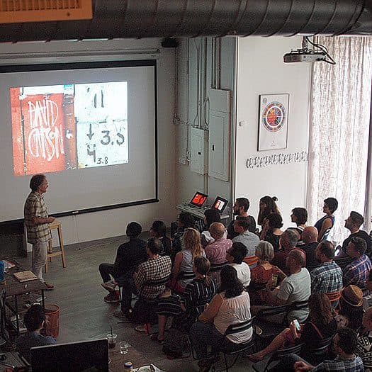 A person in a checkered shirt stands at the front of a room, presenting a slideshow to an audience seated on chairs and the floor. The projected slide shows a red background with white text. Various electronic devices are visible around the room.