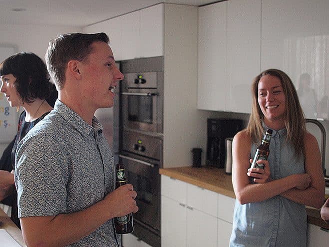 Three people are socializing in a modern kitchen. A man holding a beer bottle is smiling and talking to a woman, who is also holding a beer bottle and smiling. A third person stands in the background, slightly turned away. The kitchen is well-lit with white cabinets.