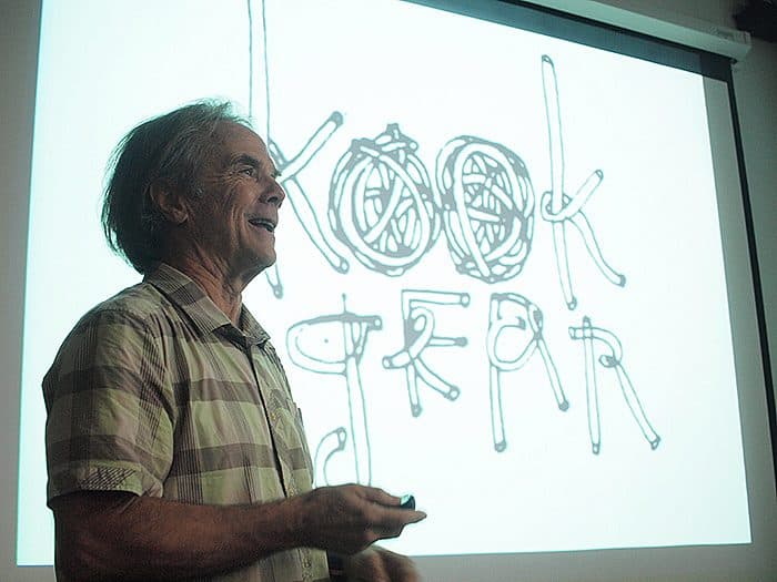 A man stands in front of a screen giving a presentation. The screen displays the text "Kook Gear" drawn in a creative, twisted rope-like font. The man is smiling and appears to be speaking, holding a remote in his hand.