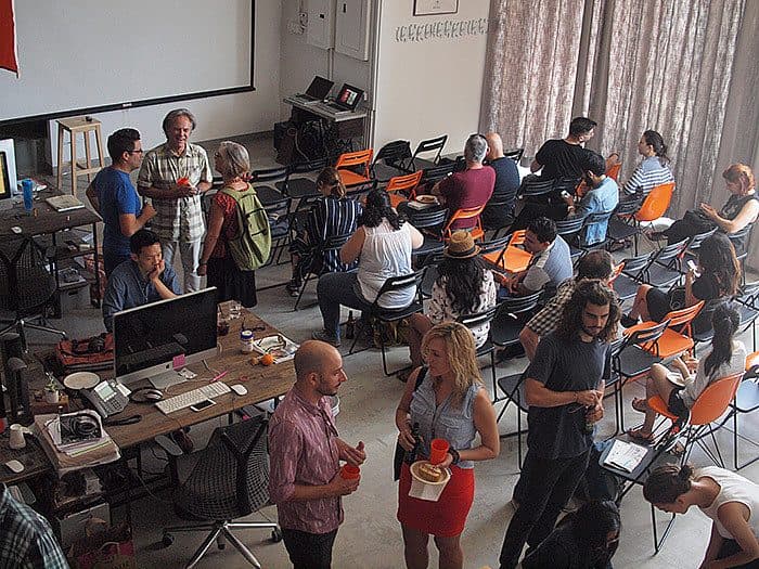 A diverse group of people are mingling in a well-lit room with rows of chairs facing a projector screen. Some individuals are seated, while others are standing and chatting. Tables with laptops and snacks are visible, and large windows with curtains provide natural light.