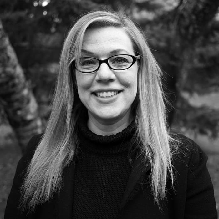 Black and white portrait of a smiling person with long, straight hair wearing glasses and a dark sweater. They are standing outdoors in front of a blurred background featuring tree trunks and foliage.