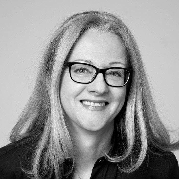 A grayscale photograph of a person with long, straight hair wearing black-framed glasses. They are smiling and appear to be dressed in a dark top. The background is plain and light in color.