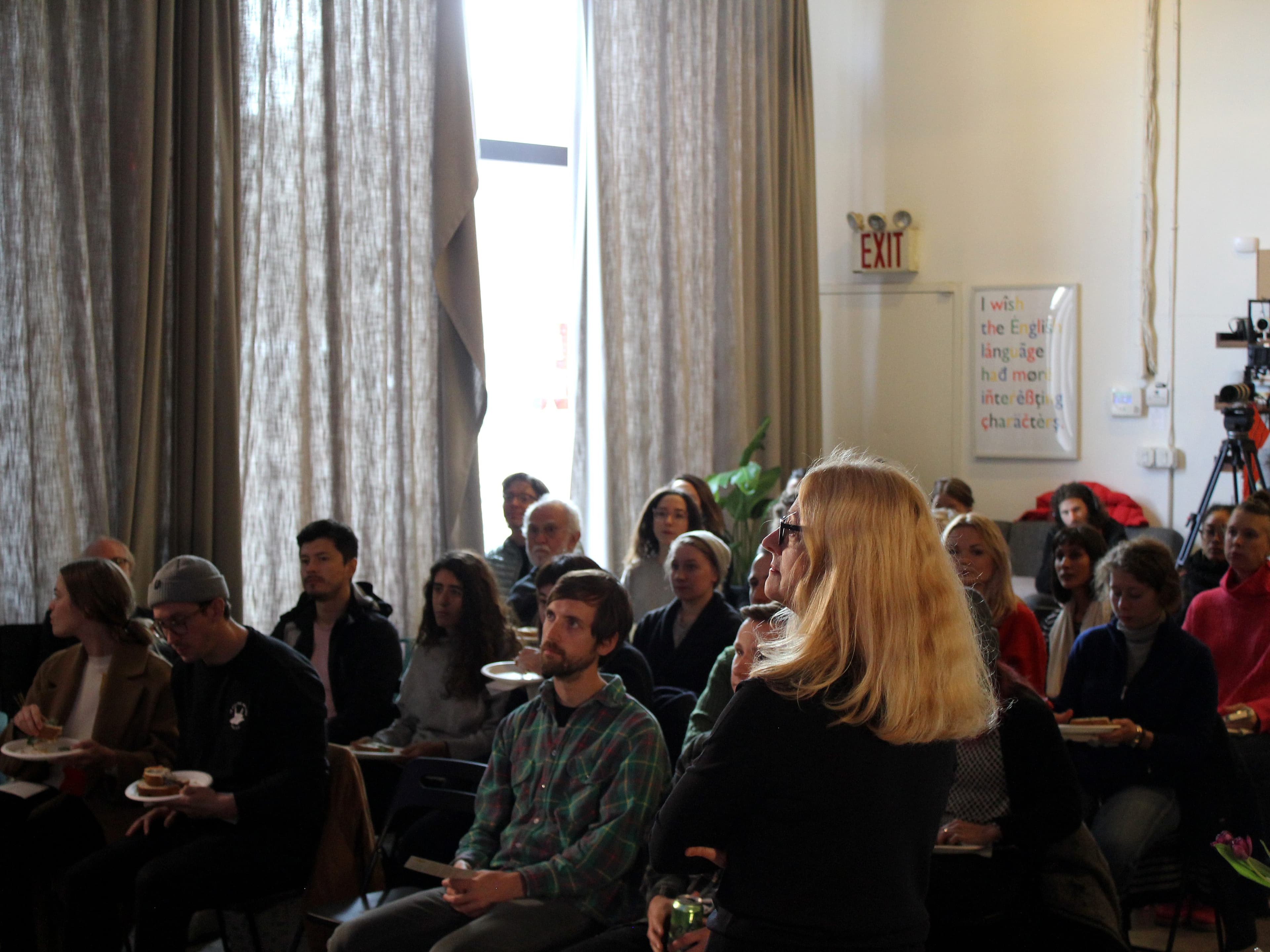 A group of people sit attentively in a room with large windows covered by curtains. A blonde woman stands in the foreground, facing the audience. Some attendees have food in their hands. There's an exit sign on the wall and a whiteboard with writing in the background.