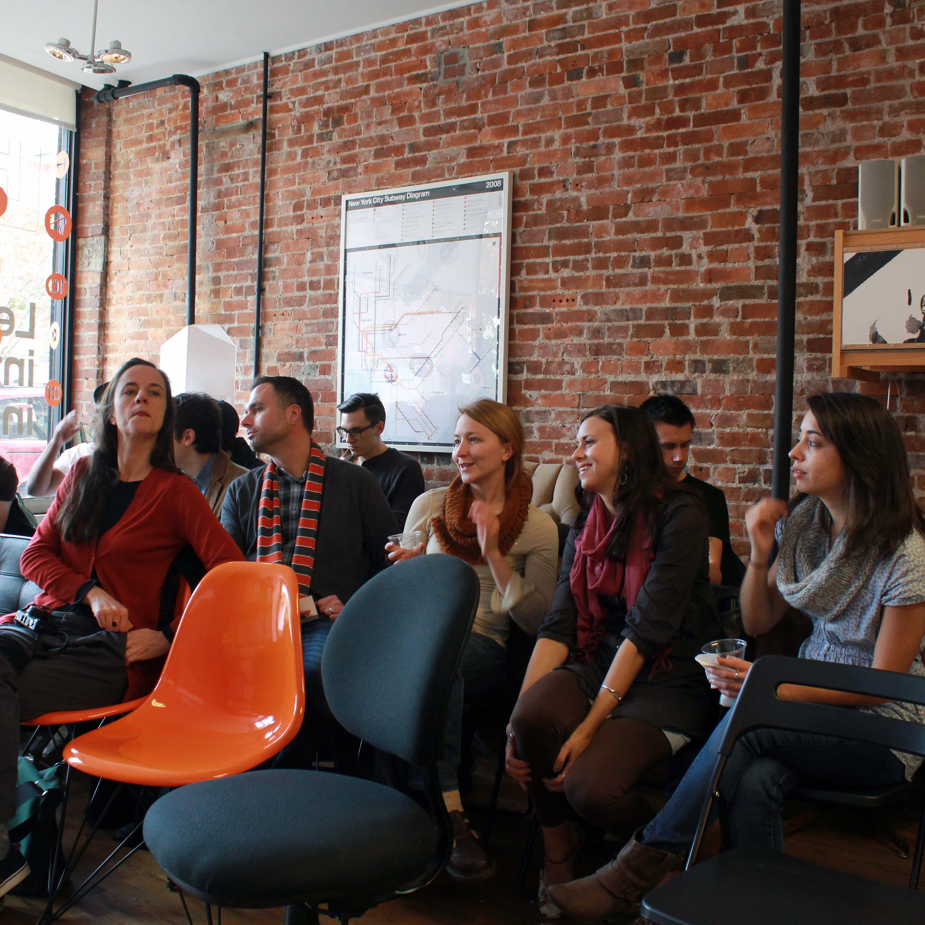 A group of people sit and chat in a casual indoor setting with brick walls and an eclectic mix of chairs. Some individuals are seated on a couch, while others are on various chairs. A map and posters decorate the wall. The atmosphere appears relaxed and social.