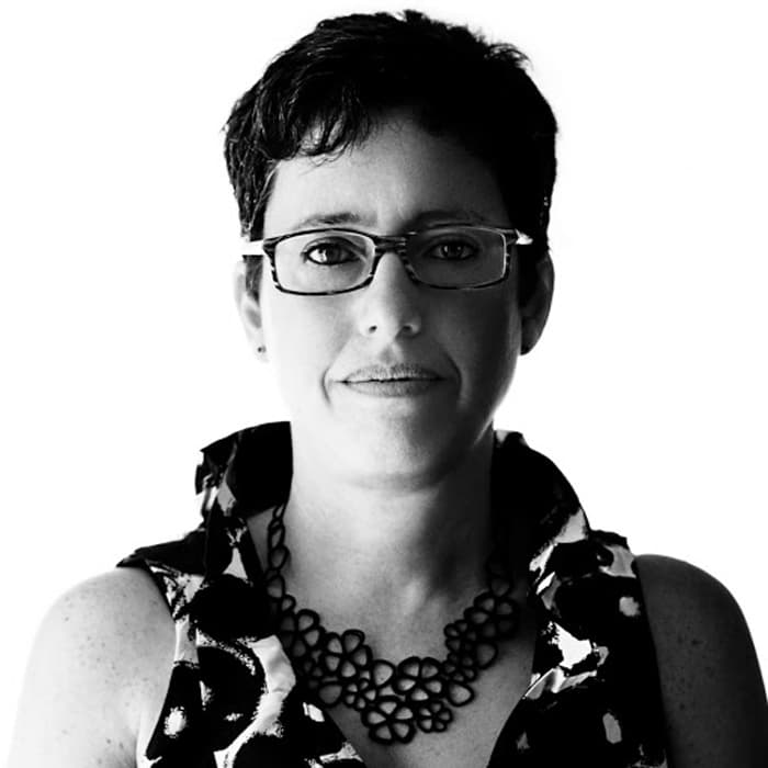 Black and white portrait of a person with short hair, glasses, and a sleeveless patterned top. The person is facing the camera with a neutral expression. The background is plain white, creating a high contrast with the subject.