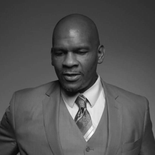 Black and white photo of a man wearing a three-piece suit and a patterned tie. He has a bald head and a trimmed beard, and he appears to be looking slightly downward with a neutral expression.