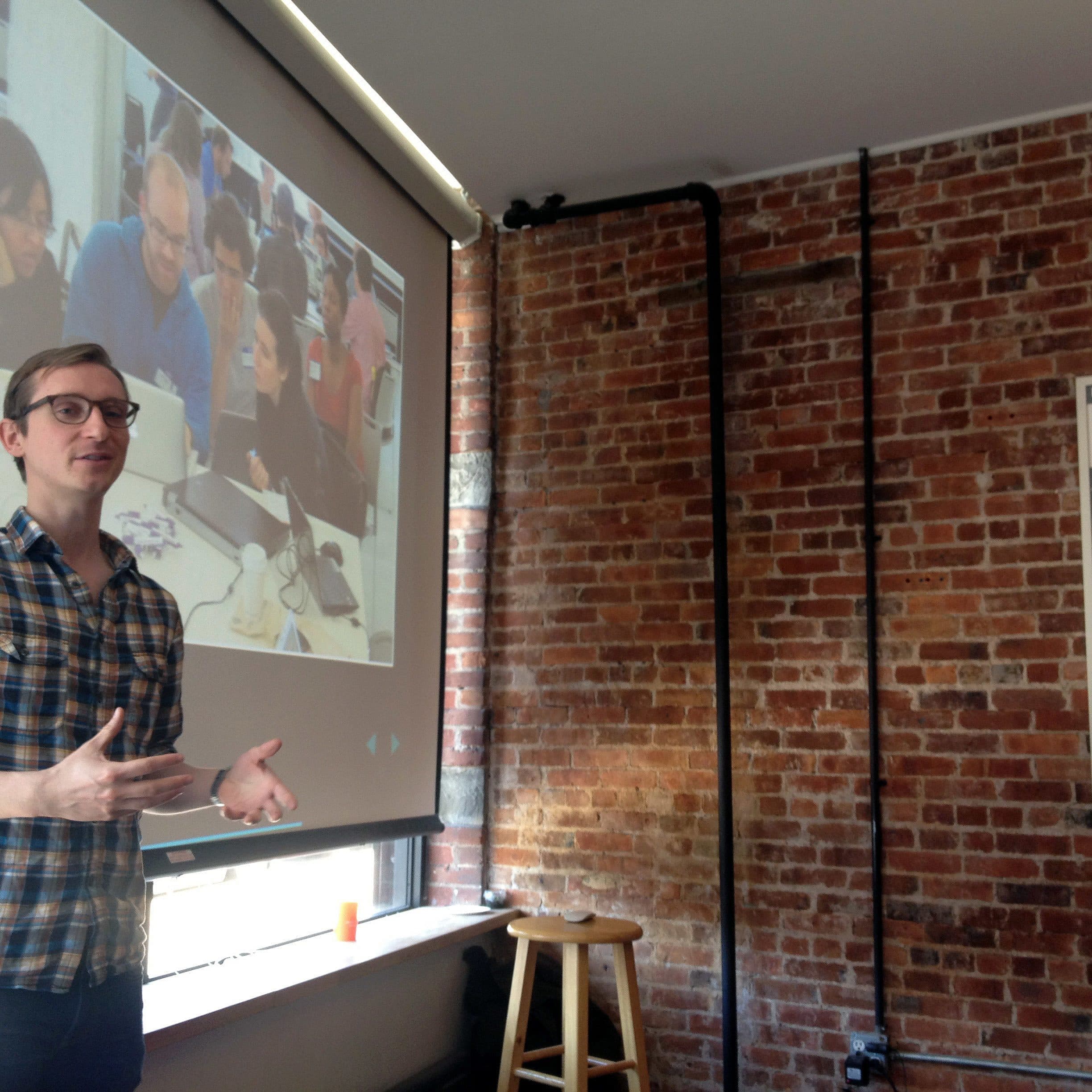 A person wearing glasses and a plaid shirt stands in front of a screen, gesturing with hands. The screen displays a photo of people at a table with laptops. The setting has a brick wall and a New York City subway map. A wooden stool is nearby.