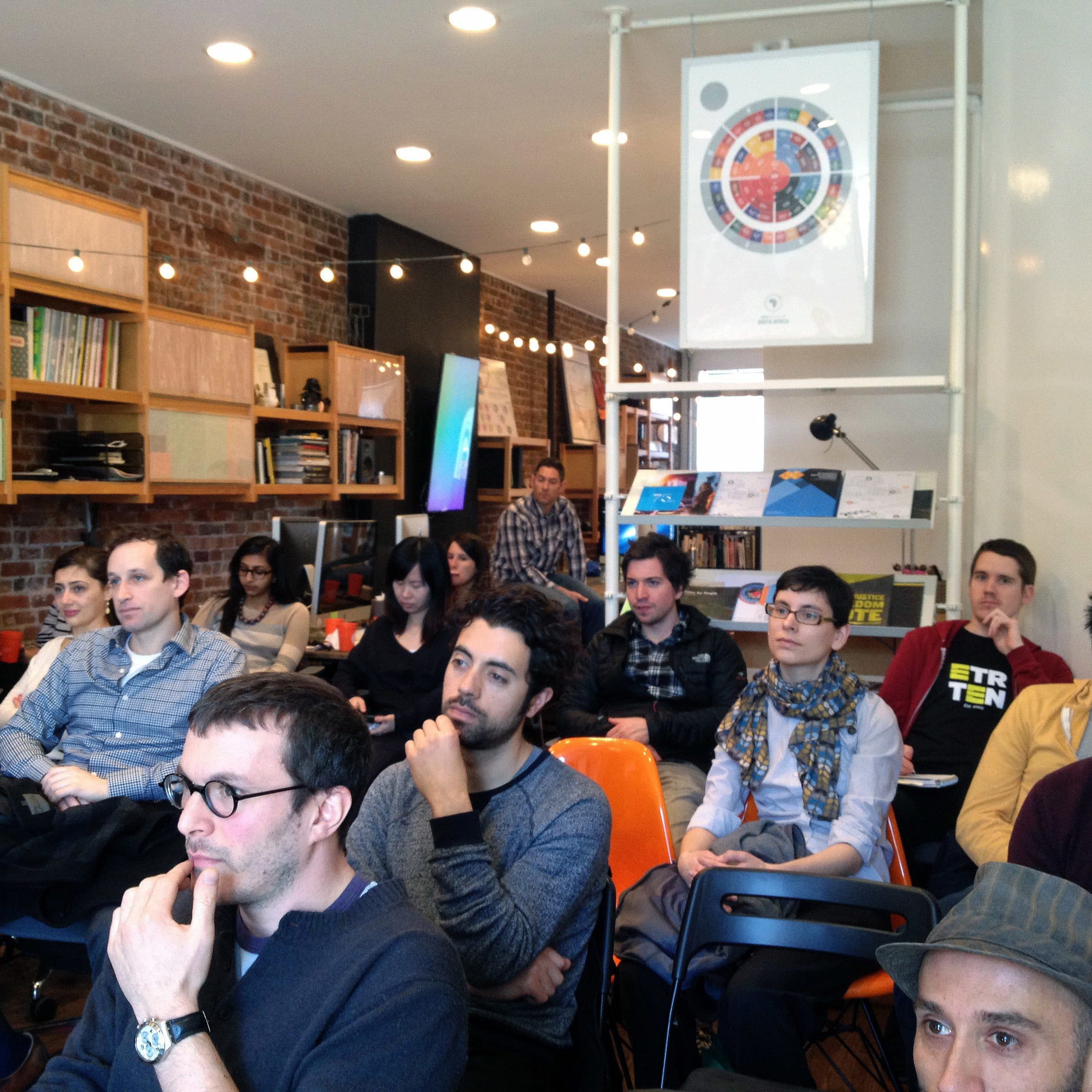 A diverse group of people sits attentively in a well-lit room with brick walls and shelves filled with books. They are facing forward, possibly listening to a presentation or lecture. A colorful chart is displayed on the wall behind them.