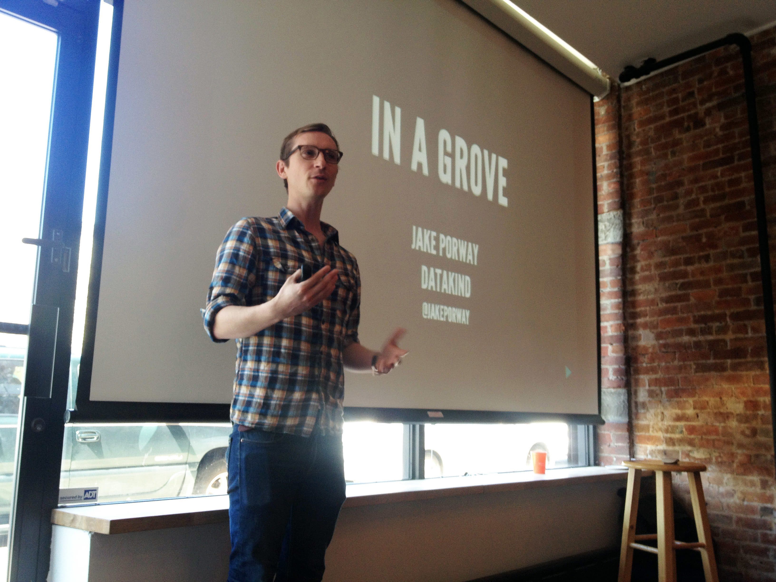 A man wearing glasses and a plaid shirt speaks in front of a projected screen displaying the title "In a Grove," along with the names "Jake Porway" and "DataKind." A wooden stool is placed near the brick wall to his right.