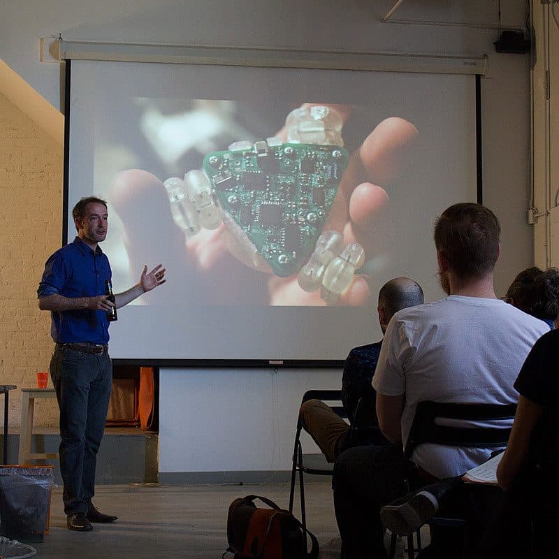 A man wearing a blue shirt is giving a presentation to a seated audience. He is pointing to a large projection of an electronic circuit board on a screen behind him. The room is dimly lit, with various equipment and chairs visible.