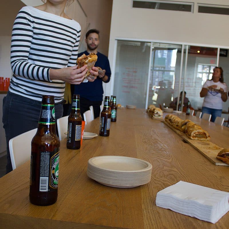 A group of people are gathered around a wooden table in a modern indoor space. On the table, there are bottles of beer, a stack of napkins, and a long serving board with bread and pastries. Several individuals are holding plates of food, engaging in casual conversation.