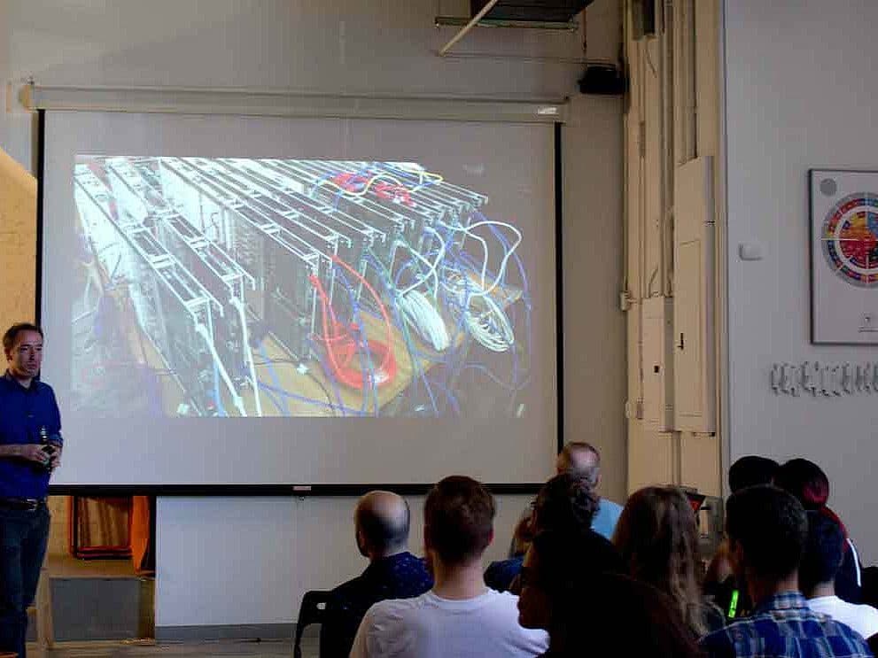 A man gives a presentation to a seated audience in a room with white walls. The projector screen behind him displays an image of a complex arrangement of wires and hardware components. A colorful poster is mounted on the wall to the right of the screen.