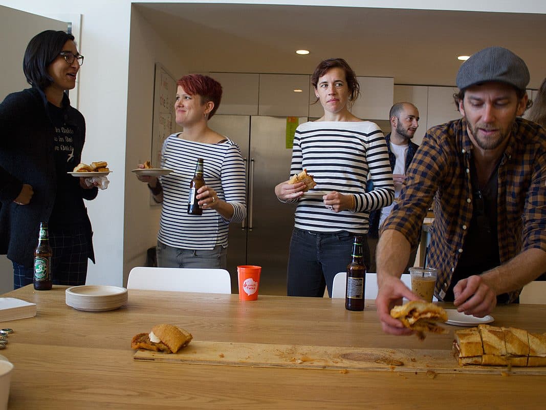 A group of people gather in a kitchen, enjoying sandwiches and drinks. One person in a hat is reaching for food on a wooden table, while others stand and chat. There are drinks and food items on the counter in the background.