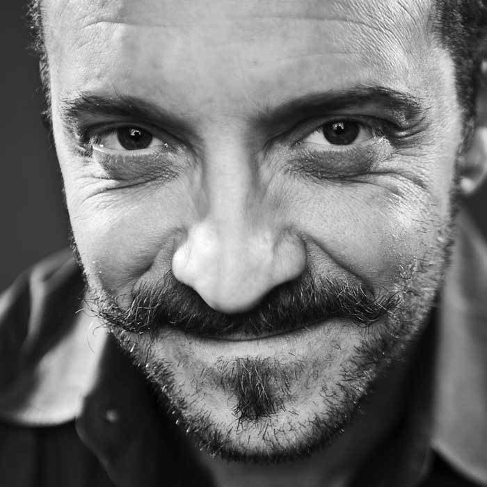 Close-up black-and-white image of a man's face with a slightly mischievous expression. He has a mustache and beard with some wrinkles visible around his eyes, which are looking directly at the camera. The background is blurred, drawing focus to his facial features.