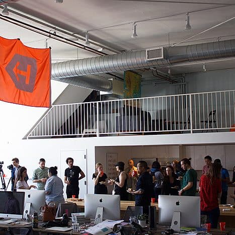 A spacious office with an open floor plan features several desks equipped with computers in the foreground. In the background, a group of people stand and converse near a snack table. A large orange flag with a geometric logo hangs from the ceiling.