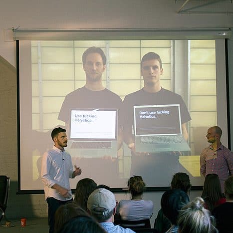 Two men stand in front of a seated audience, presenting a slide with side-by-side photos. One man holds a laptop displaying "Use fucking Helvetica," and the other holds a laptop with "Don't use fucking Helvetica." The presenters discuss typography passionately.