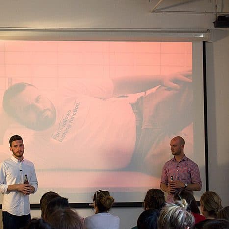 Two men stand in front of an audience giving a presentation. They are holding bottles and standing on either side of a projected image that shows a man lying down and wearing a T-shirt. The audience watches intently, seated in rows facing the presenters.