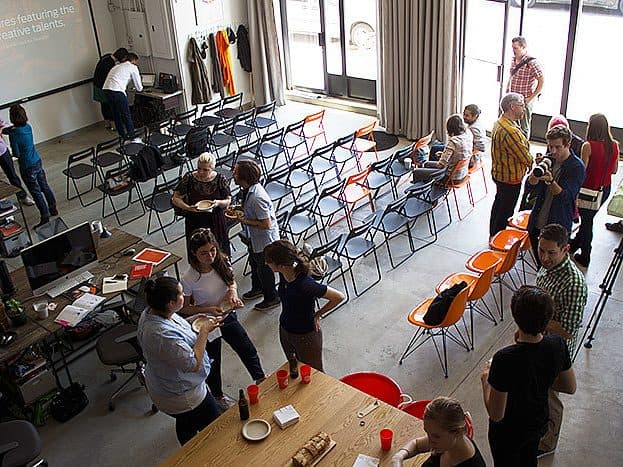 An overhead view of a workshop setup in a spacious room. Several people are scattered throughout, engaging in conversations. Rows of black and orange chairs are arranged facing a projector screen. Tables with various materials and refreshments are also visible.