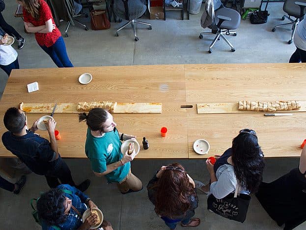 A group of people gather around a long, rectangular wooden table. They are standing and appear to be eating and socializing. On the table are plates, cups, and a long serving board with sliced bread. Additional chairs and desks can be seen in the background.
