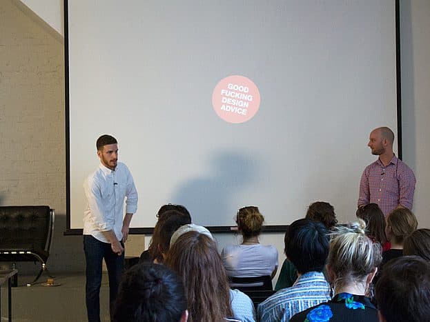 Two men stand at the front of a classroom addressing an audience. One man wears a white shirt, and the other wears a red checkered shirt. The projection screen behind them displays a circular text that reads "GOOD FUCKING DESIGN ADVICE.
