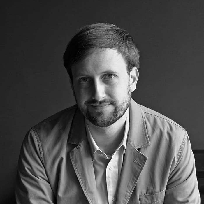 Black and white portrait of a person with short hair and a beard. They are wearing a button-up shirt and a blazer. They are looking at the camera with a subtle smile and seated against a plain, dark background.