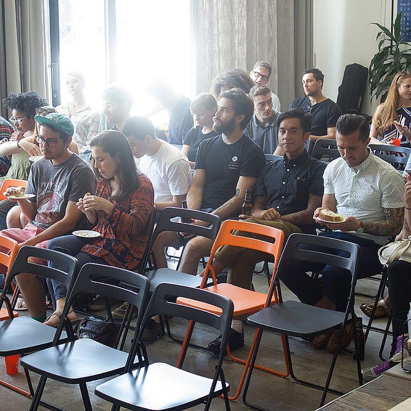 A group of people sits in rows of colorful chairs, attending an indoor event. Some people are eating or holding plates, while others are engaged in conversation. The room is well-lit with natural light from large windows, and a few plants and equipment are in the background.