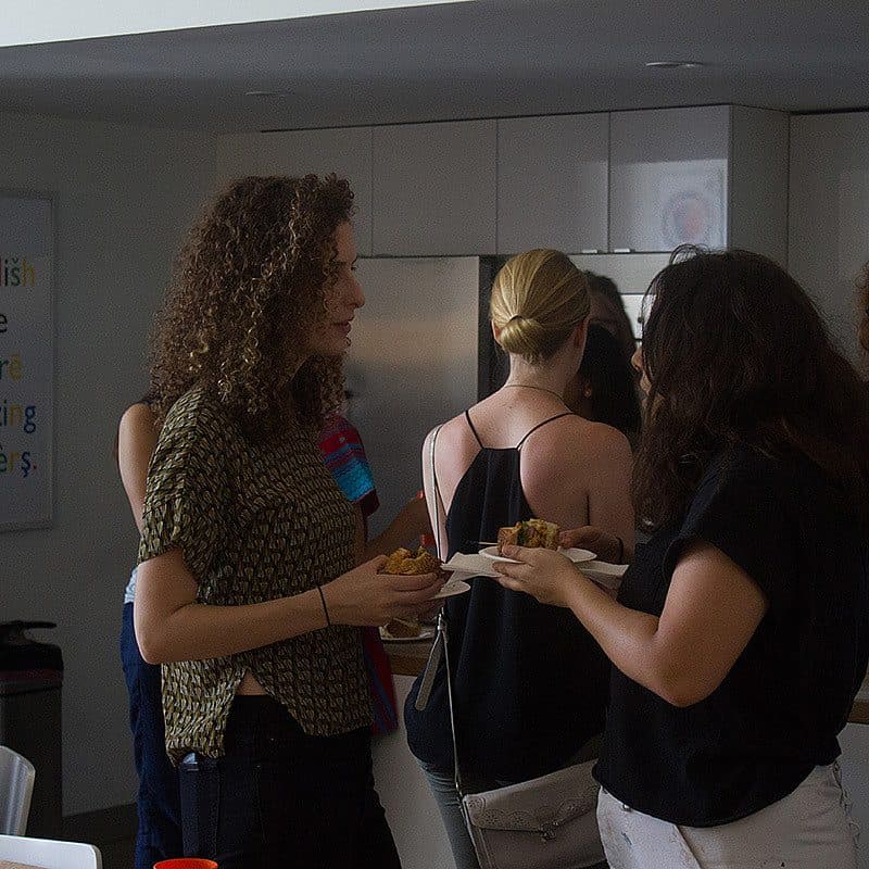 A group of people socializing in a kitchen, holding plates of food and engaged in conversation. The kitchen has white cabinets and a colorful sign on the wall that reads, "I wish the English language had more interesting characters.