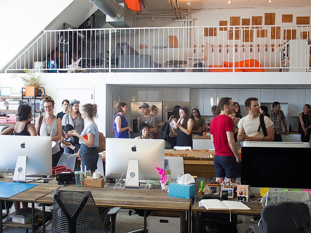 A busy office space with high ceilings and a mezzanine level. Multiple people are gathered around, some engaged in conversations, while others work at desks with computers. The room has a modern design with a mix of natural light and industrial elements.