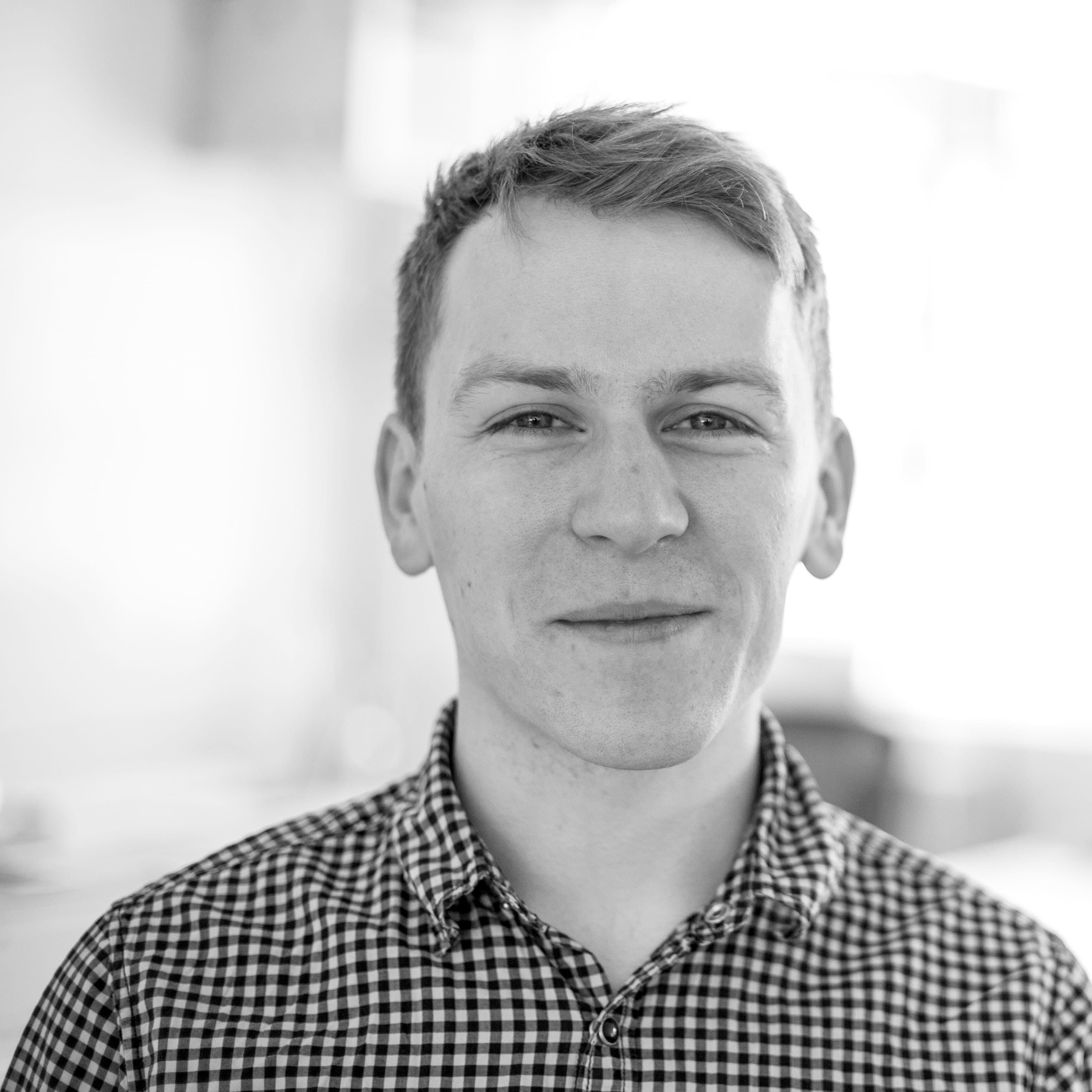 Black and white photo of a person with a slight smile, wearing a checkered shirt. The background is soft and out of focus, creating a contrast that brings attention to the subject's face.
