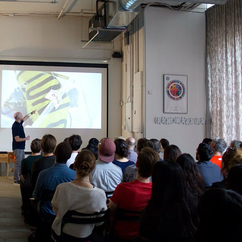 A man stands at the front of a room giving a presentation to a seated audience. He points towards a projected image of sneakers on a screen. The room has white walls, a chart on the wall, and tall curtains covering the windows.