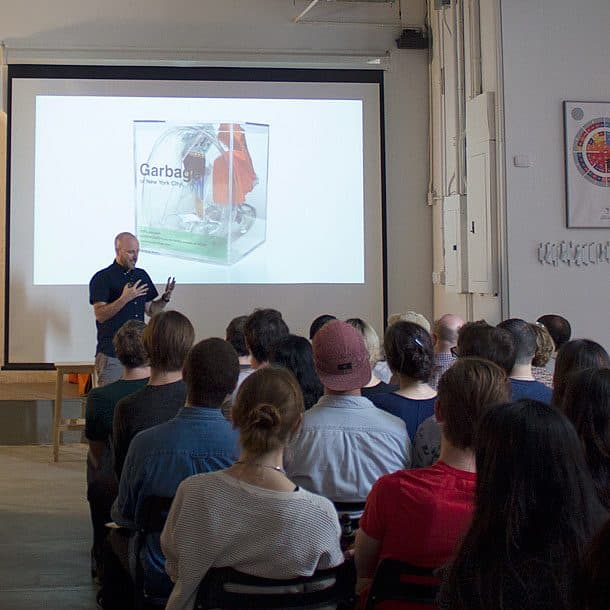 A man is giving a presentation to a seated audience in a room. He stands in front of a screen displaying an image of a project titled "Garbage." The audience attentively watches the presenter, with some people taking notes. The room has a minimalist design with high ceilings.