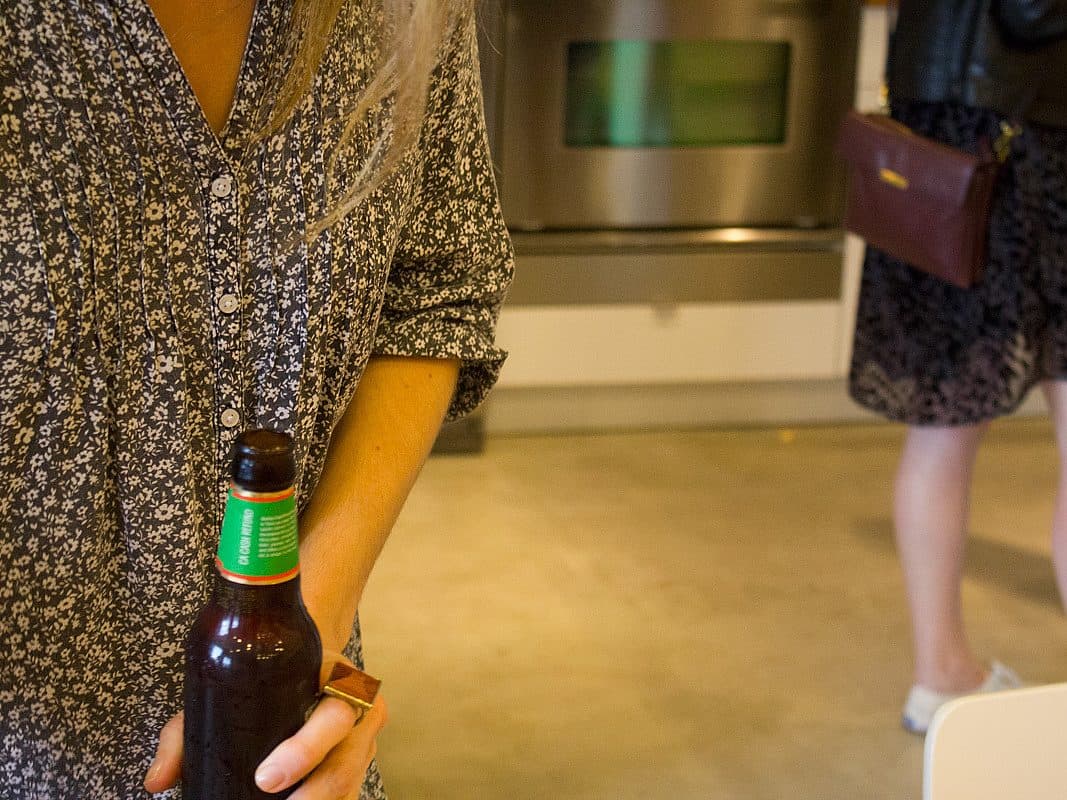 A person holding a brown glass beer bottle is partially visible, wearing a patterned shirt. In the background, another person with a patterned skirt and leather jacket stands, with a brown crossbody bag and white shoes. A stainless steel appliance is also visible.