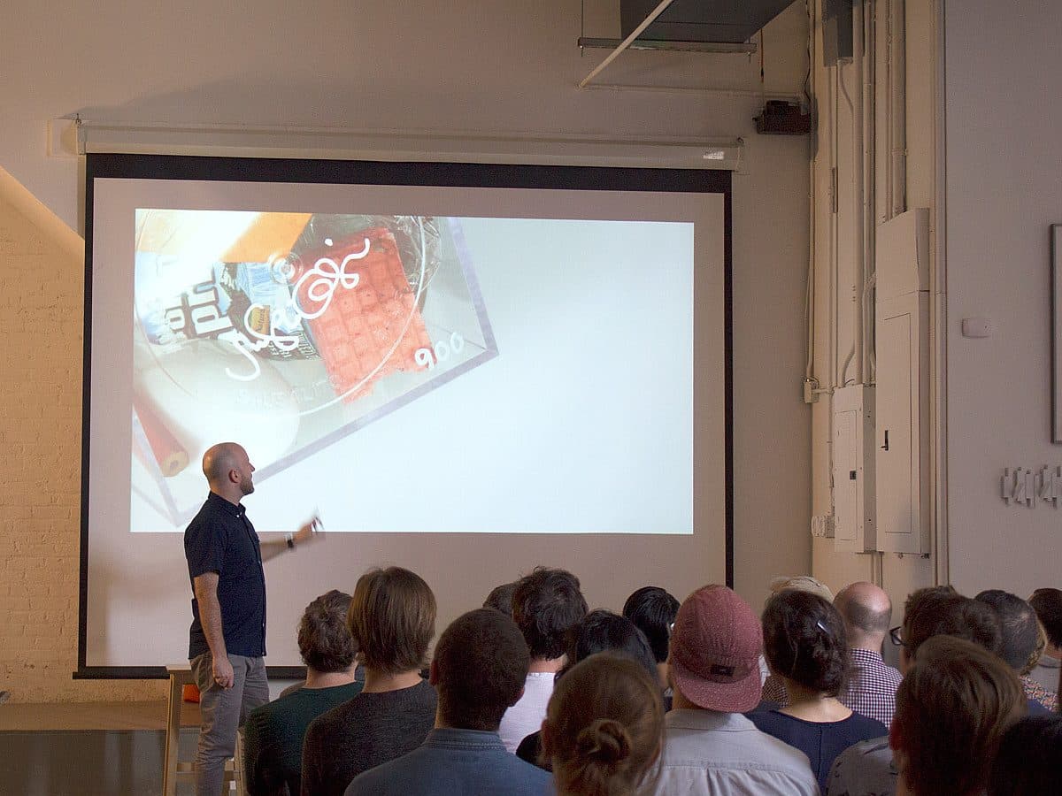 A man stands in front of an audience, pointing at a screen displaying a colorful graphic during a presentation in a modern room with white walls and industrial décor. Rows of seated individuals face the presenter, attentively listening.