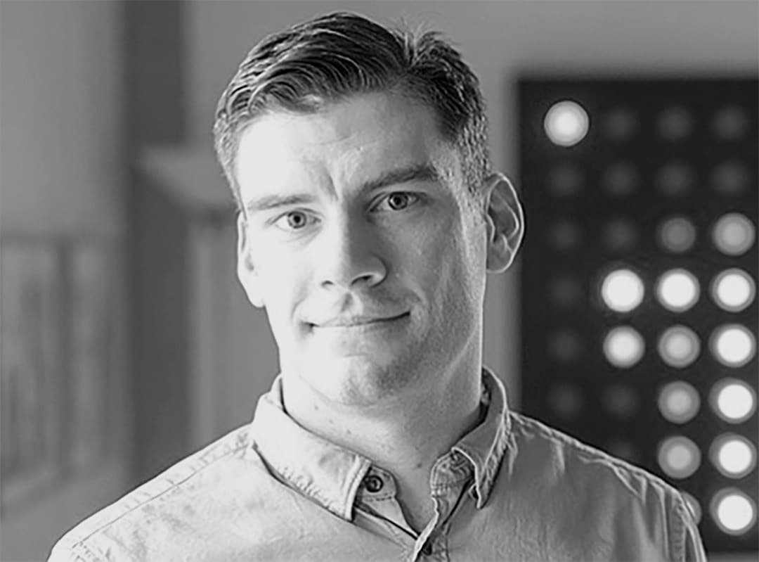 Black and white image of a person with short hair standing indoors, wearing a button-up shirt. The background is blurred with a pattern of circular lights on the right. The individual is looking straight at the camera with a neutral expression.