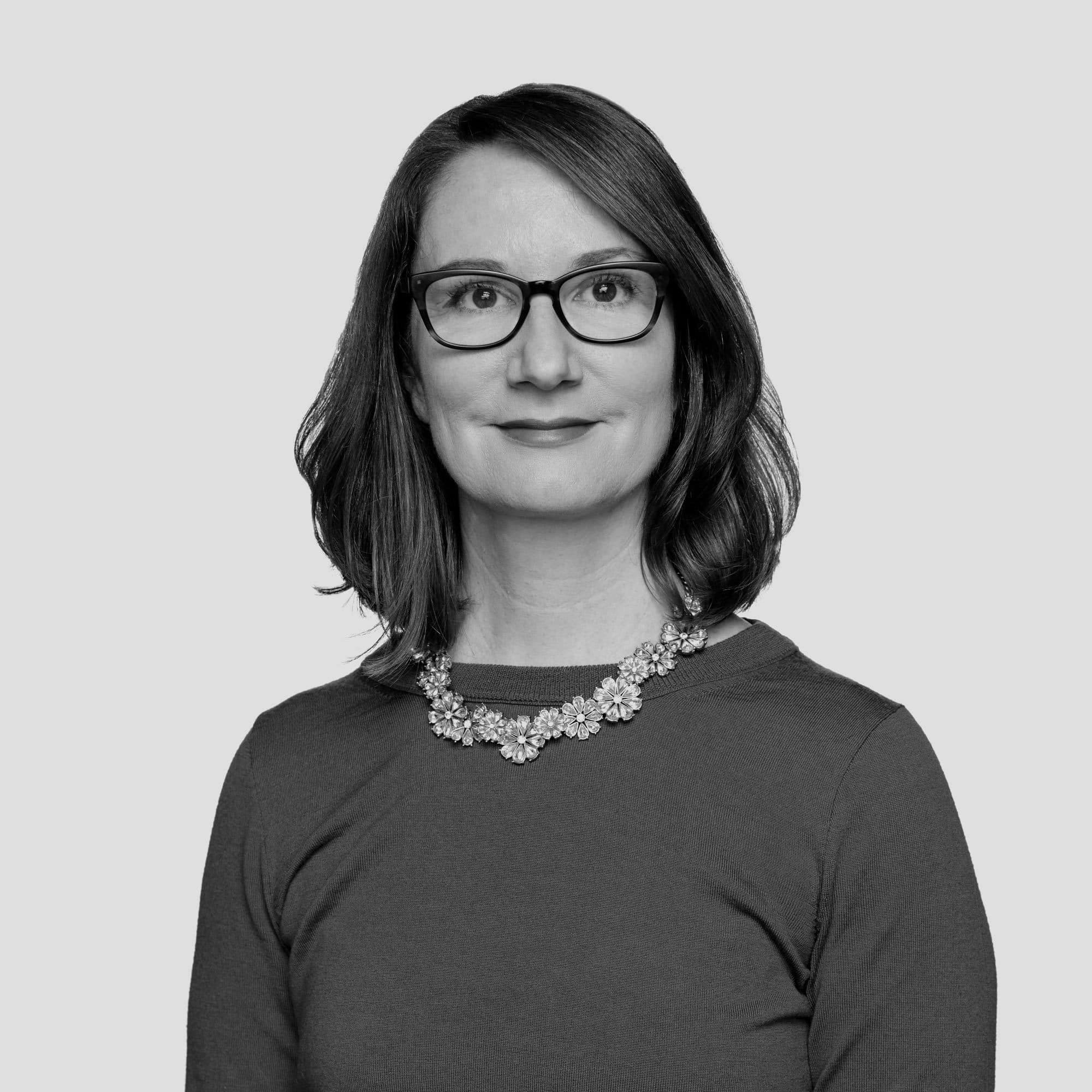 Black-and-white portrait of a person with shoulder-length hair, wearing glasses. They are dressed in a plain, long-sleeved top and a detailed floral necklace. The background is a solid light gray. The person is looking directly at the camera with a slight smile.
