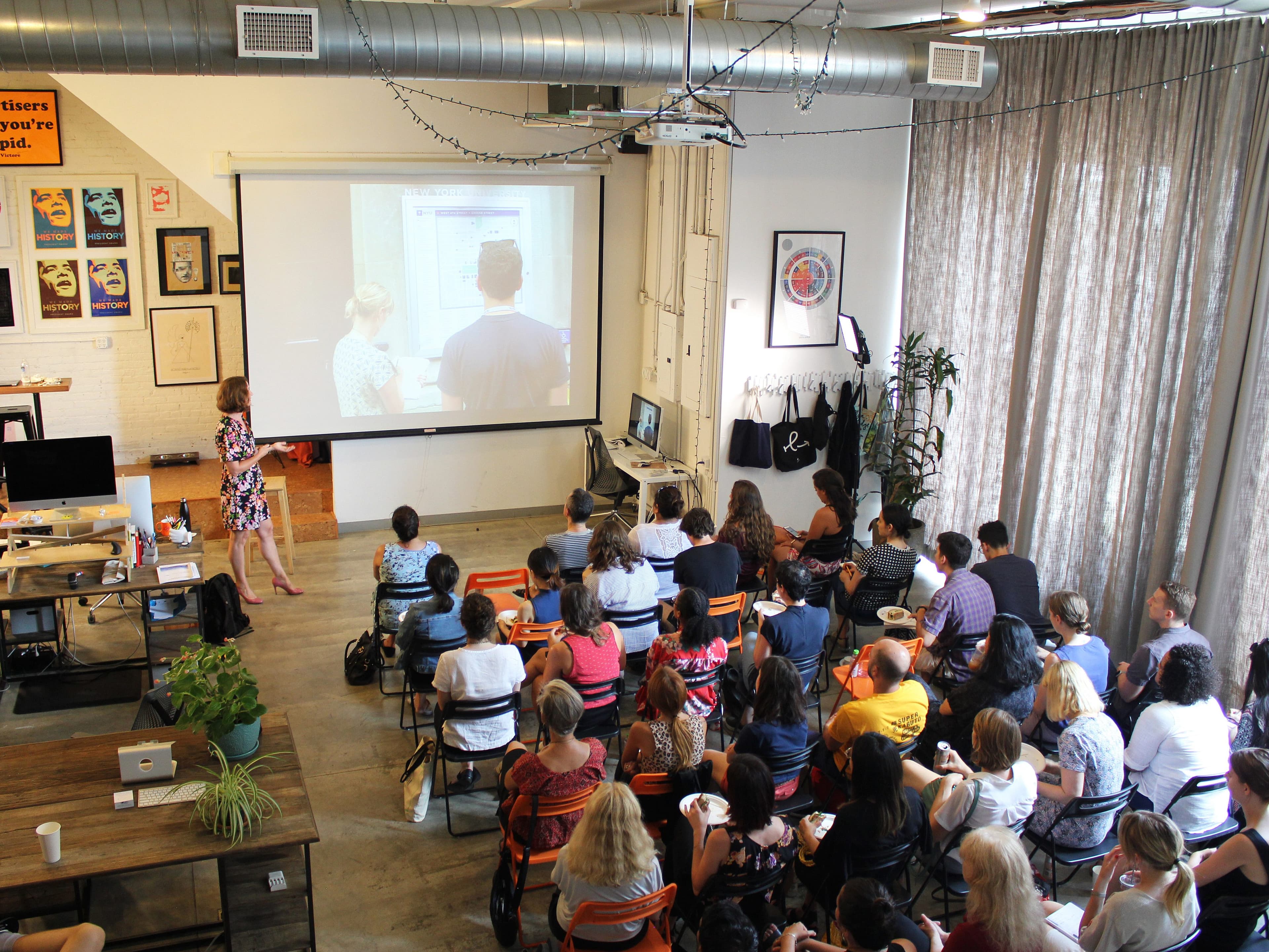 A woman in a patterned dress stands at the front of a room, presenting to a seated audience. The audience is facing a large projector screen displaying an image of two people. The room is bright, with tall windows, a mix of industrial and modern decor, and wall posters.