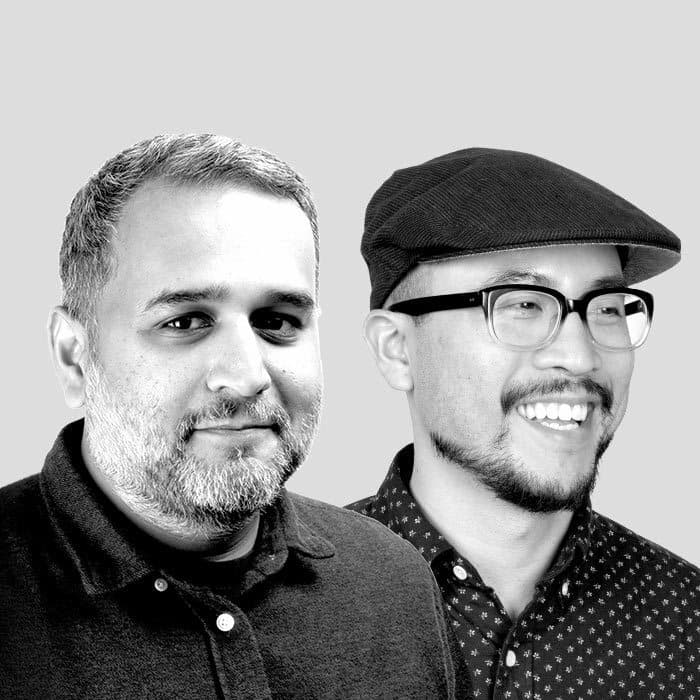 Two men are photographed side by side against a plain background. The man on the left has short, gray hair, a beard, and is wearing a dark collared shirt. The man on the right has glasses, a beard, and is smiling while wearing a flat cap and patterned shirt.