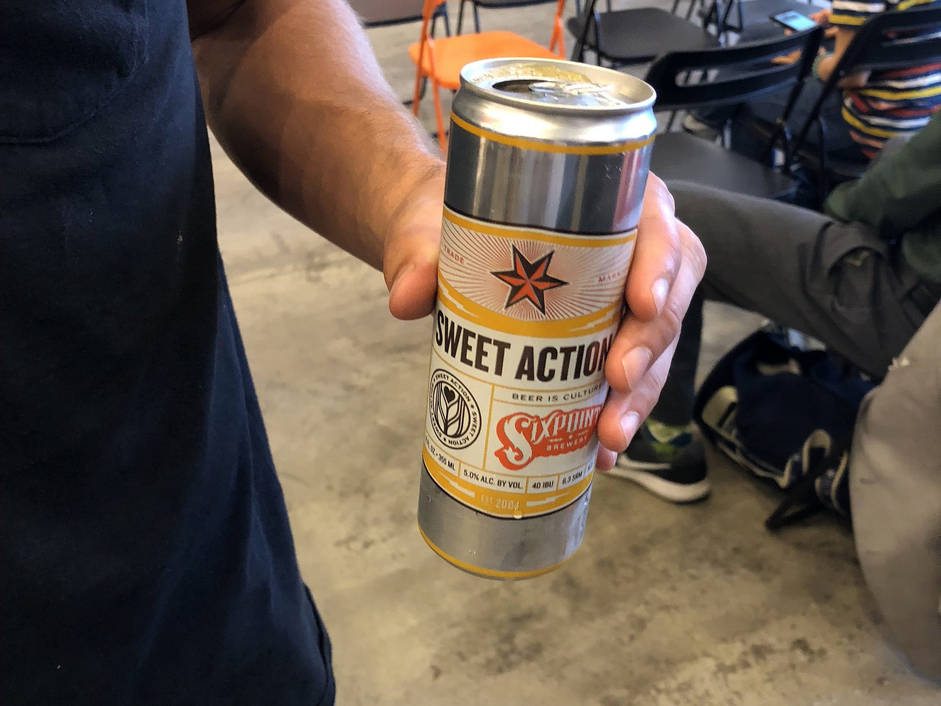 A person holding a can of "Sweet Action" beer in their right hand. The background shows several empty black chairs and one orange chair, with another individual sitting and looking down, dressed in a striped shirt. The setting appears to be a casual indoor space.