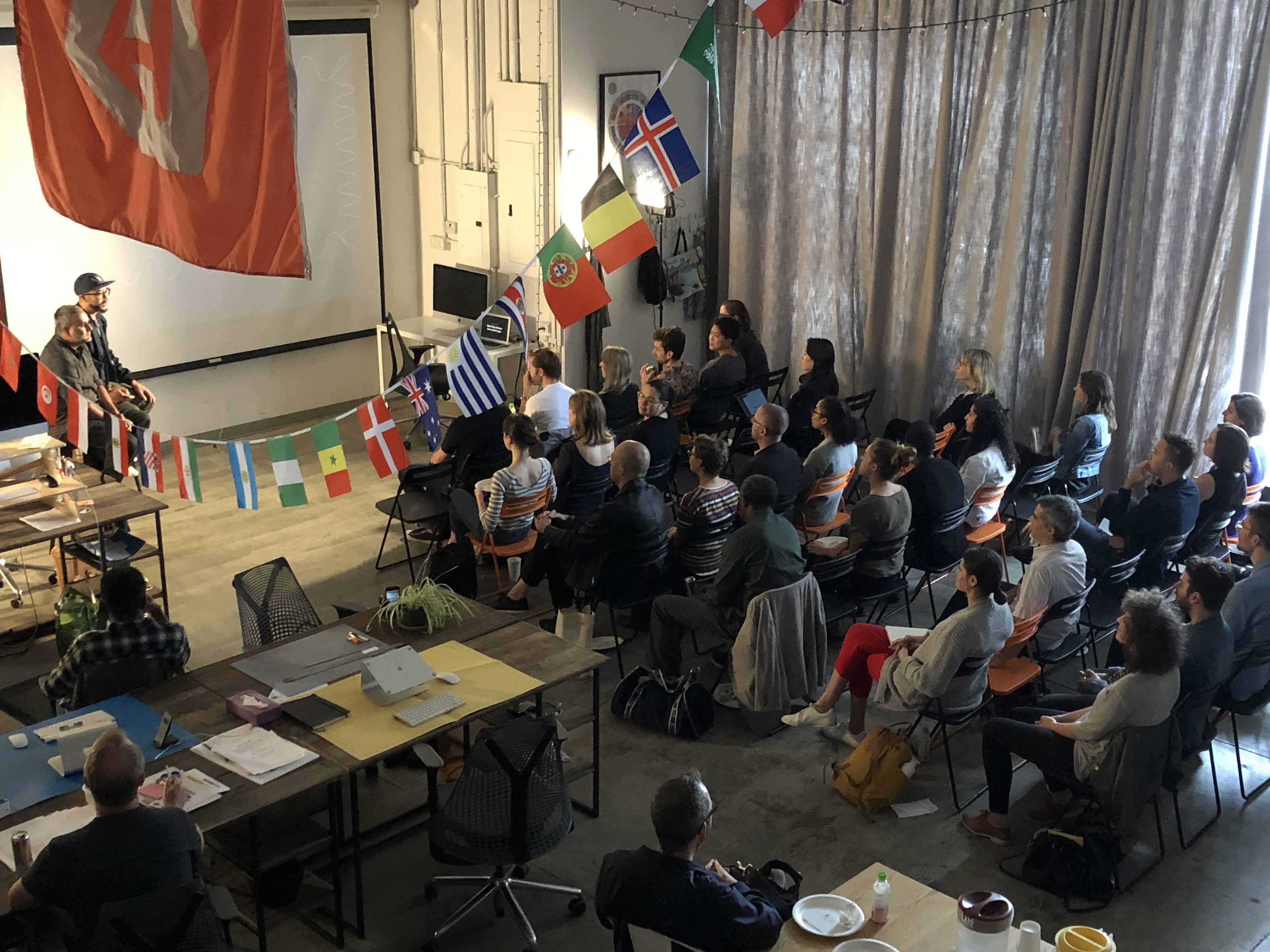 A group of people sit in a room facing a speaker at the front. The speaker is next to a large screen, and multiple international flags hang from the ceiling. Tables and chairs are arranged around the room, which has a concrete floor and high windows.