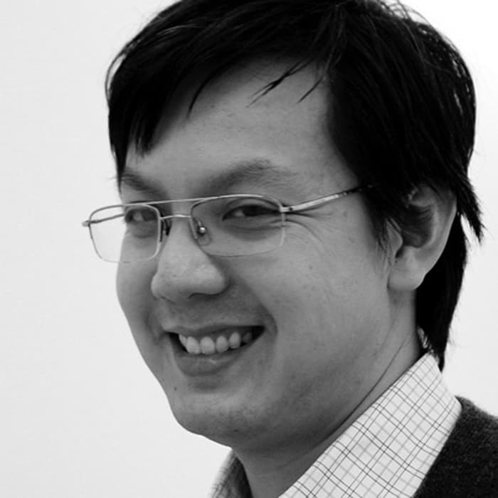 Black and white photo of a person smiling. They are wearing glasses and a checkered shirt with a sweater over it. The background is plain, and their short hair slightly covers their ear. The person is looking at the camera with a friendly expression.