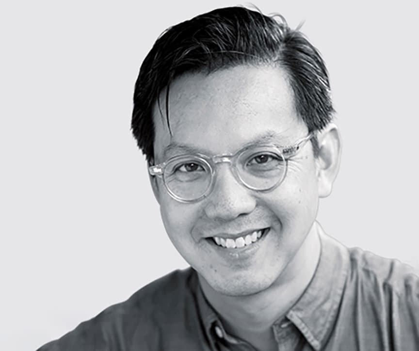 Black and white portrait of a smiling person with short, dark hair, wearing round glasses and a buttoned shirt.