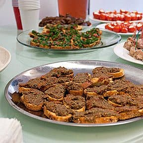 A table filled with various appetizers, including stuffed deviled eggs, toast with a spread, meat with toothpicks, and tomato-based bites. Cups and pitchers are visible in the background, suggesting a party or social gathering setting.