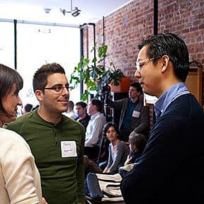 Three people are conversing in a casual setting with a brick wall in the background. A man and woman, both smiling, are facing a man with glasses who is also smiling. Other people are seated and conversing in the background. The setting appears to be a cozy cafe or meeting space.