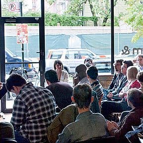 A teacher or speaker sits on a stool in front of a group of people in a casual indoor setting. The audience is seated and attentively listening. The room has large windows allowing natural light to come in. Outside, a parked car and some greenery are visible.