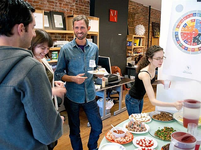 A group of four people are gathered around a table with a variety of foods. One person is serving food while the others engage in conversation. Behind them are workstations, bookshelves, and a colorful chart on the wall. The setting appears to be casual and friendly.