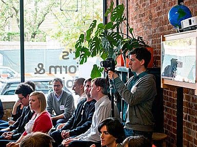 A group of people sit in a room with brick walls and large windows. A man stands holding a camera, pointing it towards the seated group. The room is decorated with plants, and a globe sits on a shelf in the background.